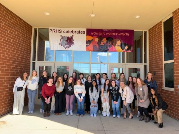 Prairie Ridge students and local leaders on the Women In Leadership Discussion Panel pose for a picture in front of the main entrance under a Womens History Month banner.
