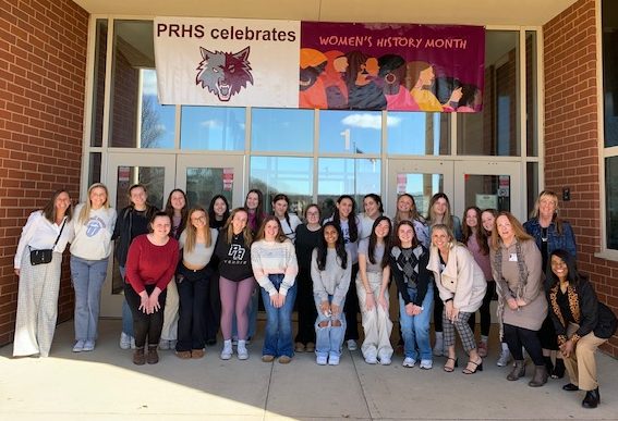 Prairie Ridge students and local leaders on the Women In Leadership Discussion Panel pose for a picture in front of the main entrance under a Womens History Month banner.